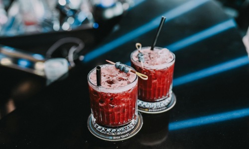 Two red berry cocktails with blueberries served on a dark bar.